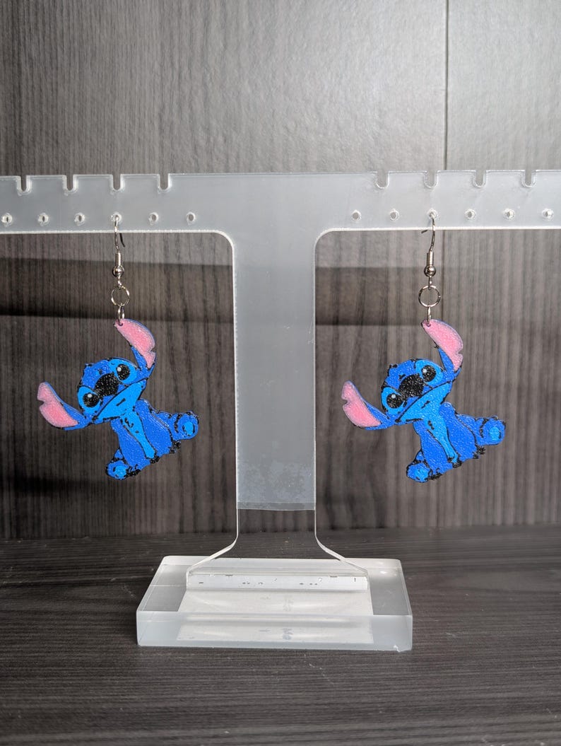 May include: A pair of blue and pink Stitch earrings. Each earring features a cartoon character design with blue body, pink ears, and black eyes. The earrings are displayed on a clear acrylic stand against a grey background.