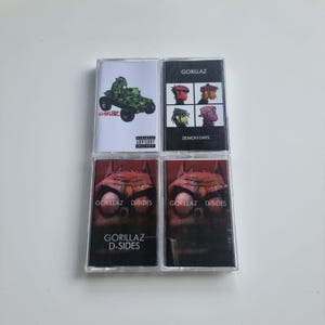 May include: Four music cassette tapes arranged in a two-by-two grid. The top left features a green vehicle illustration with the text Gorillaz and Parental Advisory. The top right shows four character portraits with the text Gorillaz Demon Days. The bottom two tapes feature a dark red creature illustration with the text Gorillaz D-Sides.