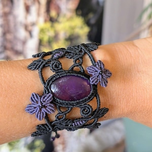 Peut inclure: Un bracelet en macramé noir avec une pierre précieuse ovale violette au centre. Le design complexe en cordon noir comprend des motifs de feuilles décoratifs et deux petites fleurs violettes à cinq pétales de chaque côté de la pierre polie.