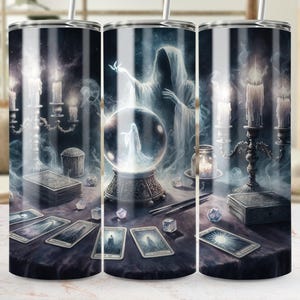May include: A tumbler featuring a mystical scene with a crystal ball, tarot cards, candles, and a spectral figure. The design is predominantly dark with shades of blue, gray, and white, creating an ethereal atmosphere. The tumbler has a silver straw.