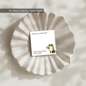 May include: A square sticky note pad with 50 sheets featuring a green frog illustration holding a coffee cup and a brown suitcase. The text reads: what's your dream job? well, in my dreams, I don't work. The note pad is centered on a white fluted dish.