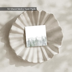 May include: A square sticky note pad with 50 sheets, featuring a watercolor illustration of a forest with green and gray pine trees and mountains at the bottom, set against a white background. The text 50 Sheet Sticky Note Pads is visible.