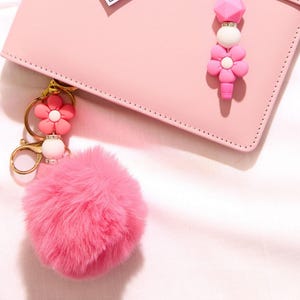 May include: A pink fluffy pom-pom keyring with a gold-coloured clasp and a pink flower charm. A pink flower charm and a beaded charm dangle from a pink leather wallet. The background is a soft pink.