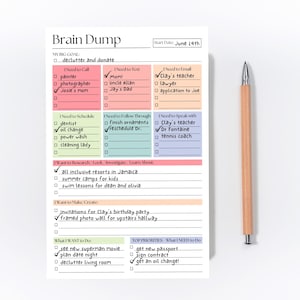 May include: A white Brain Dump notepad with a wooden pencil. The notepad has sections for goals, tasks, and notes, with the title "Brain Dump" at the top. The notepad is filled with handwritten notes and checkboxes.