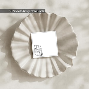 May include: A square white sticky note pad with a thin black border. The bottom left corner features a small black graphic of a stack of books with the text "IT'S A GOOD DAY TO READ" printed in a stylized black font. The pad contains 50 sheets.