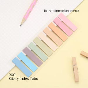 May include: A set of 200 sticky index tabs in ten trending pastel colors. The tabs are arranged in a clear plastic strip, with a pink pencil and a wooden clothespin visible in the background. Text reads: "10 trending colors per set".