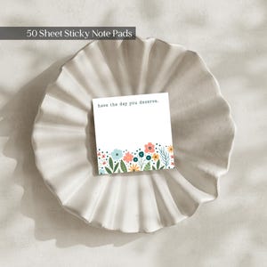May include: A square sticky note pad with 50 sheets, featuring a white background and a colourful floral border at the bottom. The text "have the day you deserve." is printed in black at the top. The design includes small flowers and leaves.