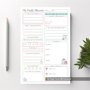 May include: A white notepad with the text "My Daily Bloom" and sections for daily planning, including intentions, gratitude, mood, sleep, and nutrition. Includes a pencil and a small plant.