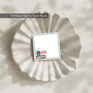 Op de afbeelding: Een vierkant memoblok met 50 vellen, voorzien van een blauwe rand en een afbeelding in de linkerbenedenhoek van een blauwe mok met een rood hart, dampend, rustend op twee boeken met de rode tekst "BOOK NERD" naast de mok gedrukt.