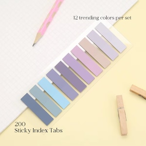 Könnte beinhalten: Ein Set aus 200 selbstklebenden Index-Tabs in 12 Trendfarben, angeordnet in einem Farbverlauf von Dunkelblau bis Hellviolett und Beige. Die Tabs befinden sich auf einer transparenten Trägerfolie, ergänzt durch einen rosa Stift und zwei Holzklammern.