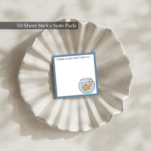 May include: A square sticky note pad with a blue border, featuring the text 'i forget so you don't have to...' and a graphic of an orange goldfish in a bowl with a small sign that reads 'WHAT WILL I DOING?'. The pad contains 50 sheets.