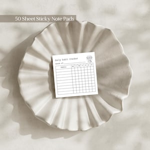 May include: A white sticky note with a habit tracker design sits on a decorative, off-white, ruffled plate. The note includes the text "daily habit tracker" and a space to track habits for the week. The top left corner of the image reads "50 Sheet Sticky Note Pads."