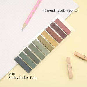 May include: A set of 200 sticky index tabs in 10 trending colors, arranged in a row. The tabs are in shades of gray, green, tan, and brown. The text "10 trending colors per set" and "200 Sticky Index Tabs" are visible. A pink pencil and two wooden clothespins are also in the image.