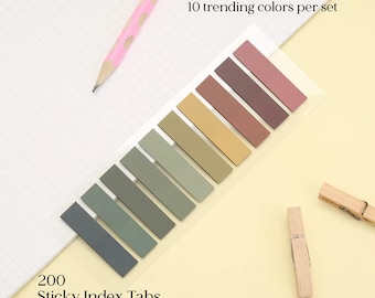 Könnte beinhalten: Ein Set mit 200 Haftnotiz-Registerkarten in 10 Trendfarben, in einer Reihe angeordnet. Die Registerkarten sind in Grau-, Grün-, Beige- und Brauntönen gehalten. Der Text "10 Trendfarben pro Set" und "200 Sticky Index Tabs" sind sichtbar. Ein rosa Bleistift und zwei Wäscheklammern aus Holz sind ebenfalls im Bild.