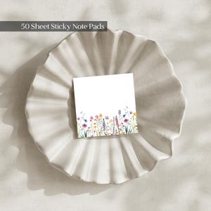 May include: A square 50 sheet sticky note pad featuring a white background with a colourful watercolour illustration of various wildflowers along the bottom edge, including shades of pink, yellow, blue, and purple stems and petals.
