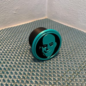May include: A teal blue and black round bottle stopper with a 3D printed design of a man's face.