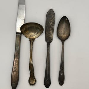 May include: An assortment of vintage silverware, including a knife, ladle, butter knife, and spoon. The silverware has a tarnished, antique appearance, with ornate detailing on the handles. The knife is stainless steel.