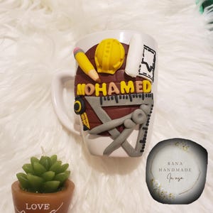 May include: White ceramic mug decorated with construction-themed elements. Features a yellow pencil, hard hat, and ruler, along with the name "MOHAMED" in gold letters. A small succulent candle and a "RANA HANDMADE" logo are also visible.