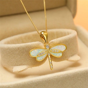 Könnte beinhalten: Eine goldfarbene Halskette mit einem Libellenanhänger. Der Anhänger hat opalisierende Flügel in Weiß und Blau, die mit kleinen, klaren Kristallen verziert sind. Der Körper der Libelle ist mit zusätzlichen Kristallen besetzt.