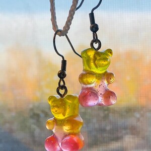 May include: A pair of gummy bear earrings with a yellow and pink ombre effect. The earrings are made of clear resin and have a shiny finish.