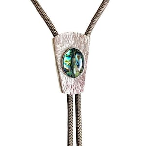 May include: A silver bolo tie with a textured, rectangular pendant. The pendant features an oval abalone shell inlay with iridescent blues, greens, and purples. The cord is a dark gray, braided material.