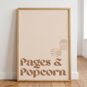 Könnte beinhalten: Ein gerahmter Kunstdruck mit hellbeigem Hintergrund und dem braunen Schriftzug "Pages & Popcorn" in einer fetten, geschwungenen Schrift. Über dem Text befinden sich einfache braune Strichzeichnungen eines Bücherstapels und einer Popcorntüte.