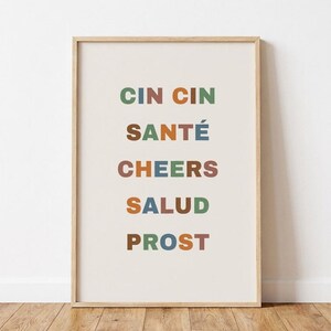 Könnte beinhalten: Ein gerahmter Kunstdruck mit dem Text CIN CIN, SANTÉ, CHEERS, SALUD und PROST in untereinander angeordneten Zeilen. Jeder Buchstabe ist in einer gedeckten Farbpalette aus Grün, Blau und Braun auf einem hellen cremefarbenen Hintergrund gehalten.