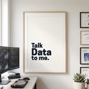 Puede incluir: Un póster enmarcado con el texto "Talk Data to me" en azul oscuro. El póster está en un marco de madera clara y cuelga de una pared blanca. El entorno de la oficina incluye monitores de computadora, un teclado y certificados enmarcados.