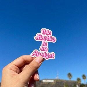 Op de afbeelding: Een gestanste vinylsticker met de tekst "this Barbie is an Analyst" in een vet, felroze, retro-geïnspireerd lettertype met een witte omlijning. De sticker heeft een geschulpte, wolkachtige vorm die de contouren van de letters volgt.