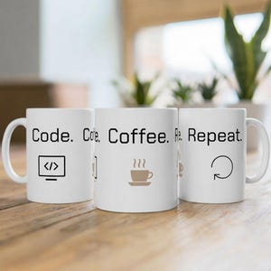 Könnte beinhalten: Drei weiße Keramikbecher mit schwarzem Text und Grafiken. Die Becher lesen "Code. Coffee. Repeat." und zeigen ein Computerbildschirm-Symbol, eine Kaffeetasse und einen kreisförmigen Pfeil. Die Becher stehen auf einer Holzoberfläche.