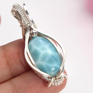 May include: A wire-wrapped pendant featuring a smooth, oval-shaped light blue Larimar gemstone with white marbled patterns. The stone is encased in a polished silver-colored metal wire cage that forms a decorative bail at the top and a small coil at the base.