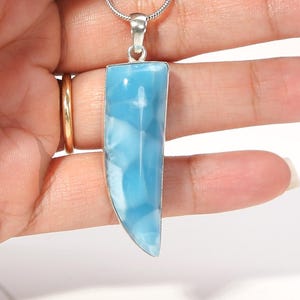 Puede incluir: Un colgante con una piedra Larimar pulida en forma de diente con patrones jaspeados en azul claro y azul cielo, engastada en un marco de metal plateado y unida a una cadena de serpiente plateada.