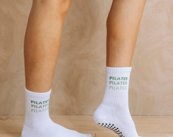 May include: A pair of white crew-length socks featuring the word PILATES printed in a repeating green font on the ribbed cuffs. The soles of the socks are equipped with small black circular grip dots to provide traction during physical activity.