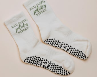 May include: A pair of white crew-length socks featuring the green text EAT, SLEEP, pilates REPEAT printed on the upper portion. The soles of the socks are covered in black circular silicone grip dots and include the text P & LOW printed in black.