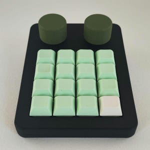 May include: A black mechanical keyboard with sixteen light green keycaps and one white keycap. Two olive green cylindrical knobs are positioned above the keycaps. The keyboard has a rectangular shape.