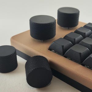 May include: A mechanical keyboard with a tan frame and black keycaps, featuring several cylindrical black knobs with a textured, knurled grip surface. Two knobs are attached to the keyboard frame, while two additional matching knobs sit nearby.