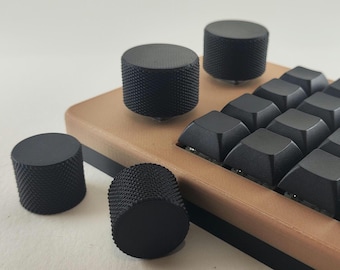 Könnte beinhalten: Eine mechanische Tastatur mit einem beigefarbenen Rahmen und schwarzen Tastenkappen, ausgestattet mit mehreren zylindrischen schwarzen Drehknöpfen mit texturierter, gerändelter Oberfläche. Zwei Knöpfe sind am Rahmen befestigt, zwei weitere liegen daneben.
