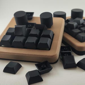 Puede incluir: Dos teclados macro personalizados con marcos de color beige impresos en 3D y teclas mecánicas negras. Cada unidad incluye dos perillas cilíndricas negras con agarre texturizado. Varias teclas negras sueltas están esparcidas en primer plano.