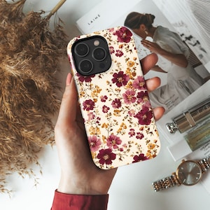 May include: A floral phone case featuring a cream background with a pattern of burgundy and yellow flowers. The case has a camera cutout and is held in a person's hand.