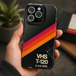 May include: Black phone case with a retro VHS tape design. The case has diagonal stripes in red, orange, and yellow. The text "VHS T-120" and "2-4-6 HRS." are printed on the case.