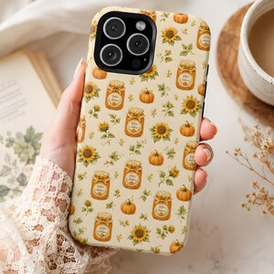 Peut inclure: Une coque de smartphone couleur crème avec un motif répétitif de citrouilles orange, de tournesols jaunes et de bocaux en verre étiquetés Golden Autumn. La coque présente une découpe noire pour l'appareil photo et une finition mate.
