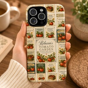 Op de afbeelding: Een beige telefoonhoesje met een collage van illustraties en tekst over tomaten. Het ontwerp bevat afbeeldingen van tomaten, tekst zoals "Rebecca's Tomato Garden" en verschillende tomatenrassen. De hoes wordt in een hand gehouden.