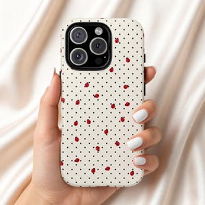 Puede incluir: Una funda de teléfono blanca con un patrón de lunares negros y mariquitas rojas. La funda tiene un recorte para un sistema de cámara de múltiples lentes. La funda del teléfono se sostiene en una mano.