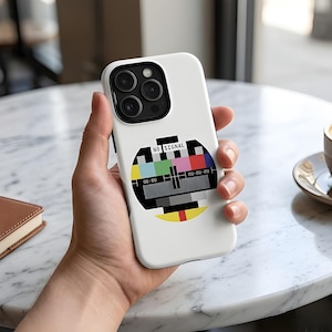 Op de afbeelding: Witte telefoonhoes met een kleurrijke "No Signal" testpatroonafbeelding. De hoes heeft een glanzende afwerking en een zwart cameralensgedeelte. De telefoonhoes wordt in de hand van een persoon gehouden.