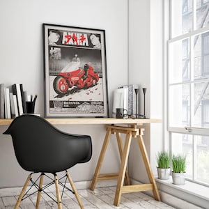 May include: Framed poster featuring a red motorcycle from the anime film "Akira." The artwork has Japanese text and illustrations of characters. The poster is displayed on a wooden desk with a black chair and a window in the background.