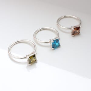May include: Three silver rings, each featuring a square gemstone in different colors: olive green, turquoise blue, and brown. The rings are displayed on a white surface, showcasing the jewelry's simple elegance.
