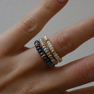 May include: Three beaded rings stacked together. One ring has dark blue pearls, another has white pearls, and the third has a mix of white and grey pearls. Gold-coloured beads separate the pearls.