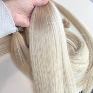 Op de afbeelding: Blonde hairextensions, gebundeld en vastgezet met zwarte elastiekjes. De extensions hebben een lichte, natuurlijke blonde kleur, met een gladde, rechte textuur. Er zijn verschillende bundels zichtbaar, met loshangende strengen.