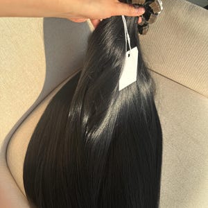 Puede incluir: Un paquete de extensiones de cabello negro. Las extensiones se sostienen en una mano y parecen lisas y rectas. La parte superior de las extensiones está asegurada con pequeñas cintas adhesivas de color claro. Una etiqueta blanca en blanco cuelga de las extensiones.