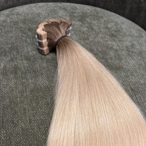 以下が含まれることがあります： 滑らかでストレートな質感のテープインヘアエクステンション。上部の接着タブ部分はダークブラウンで、そこから毛先に向かって明るいペールブロンドへと色が変化するグラデーションカラーのヘアピースです。